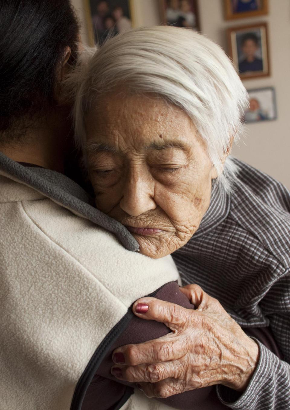 A still from the New Day film The Caretaker + The Mayor. An elderly woman with white hair hugs a person who is facing away from the camera. Her eyes are closed and her expression is vulnerable as she holds the person close. Behind them is a wall full of framed photos of a young child.