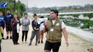 A man wearing shorts, a white t-shirt, and a vest addresses a crowd of volunteers using a megaphone at a cleanup site along the LA River. 