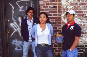 Three youth stand in front of a brick wall with grafitti. 