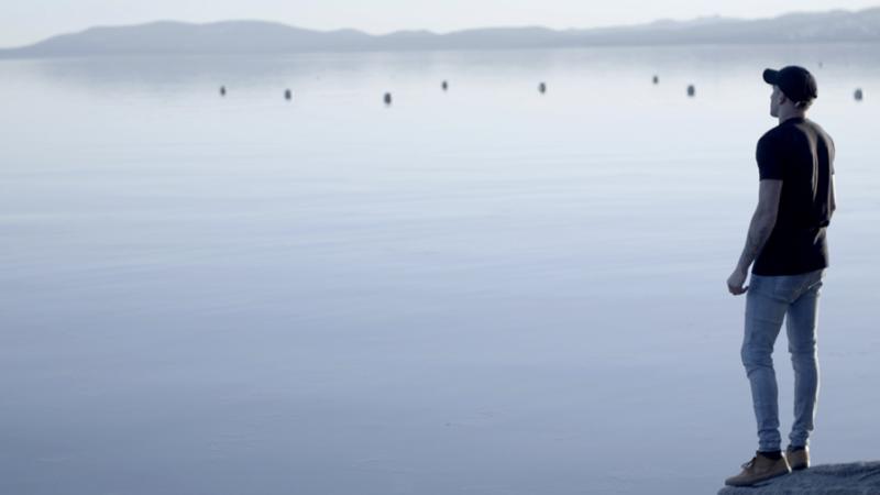 A production still from the film Momentum. A person stands to the extreme right of the frame and looks out at a lake. 