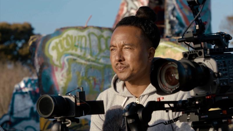 Filmmaker Adam Chan looks off to his left with pursed lips. He is surrounded by two cameras. There is graffiti on a wall in the background.