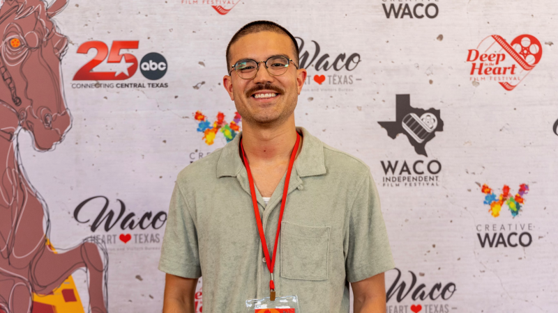 New Day Filmmaker Jake Solomon faces the camera and smiles. He wears a green shirt and is in front of a banner with logos.