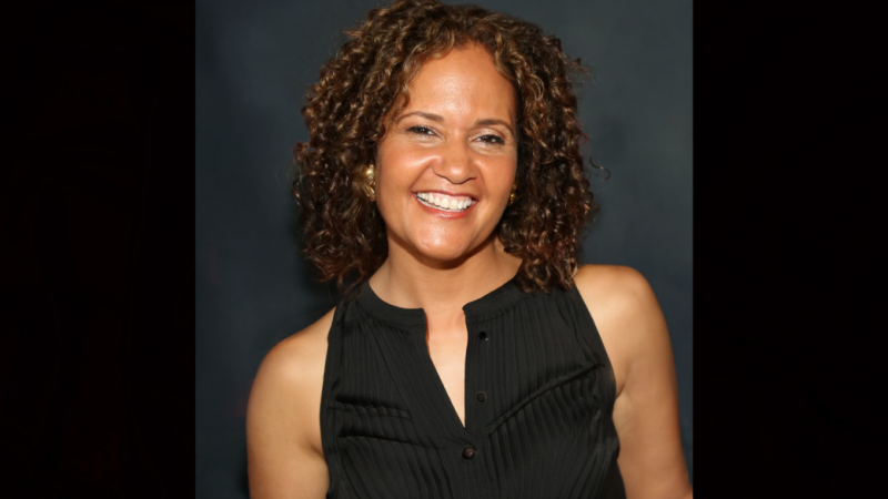 Image of a woman with dark curly hair standing in front of a navy blue background smiling broadly at the camera she wears a black tank top. 