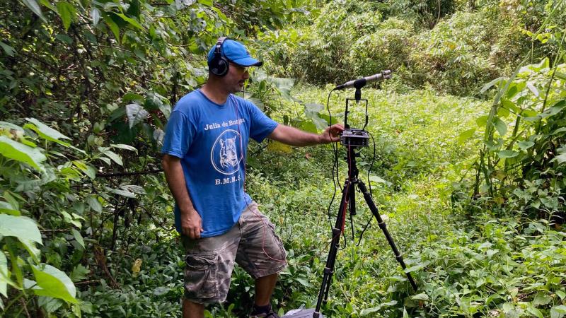 Filmmaker Juan Manuel Pagán Teitelbaum stands next to a recording device which is placed on a tripod. He is surrounded by lush greenery.