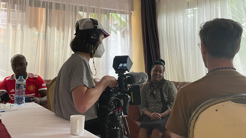 Filmmaker Trevor Zimmer is wearing a mask and pointing a camera towards a person sitting on a chair. 