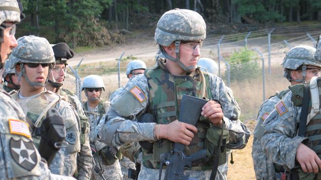 Ten men from the United States army stand in their fatigues in a field with a barbed wire fence behind them. In the center is a soldier who is looking to the right as he holds his rifle pointed downwards.