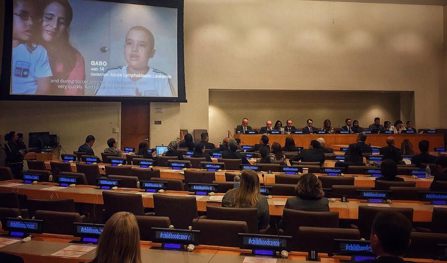 New Day Films title “How I Live” is projected on a hanging screen in a large meeting hall. On the screen, a young child sits on a woman’s lap. Gabo, a 14-year old child sits to their right. People turn to watch the film from their seats. At each seat is a digital display reading “#childhoodcancer."