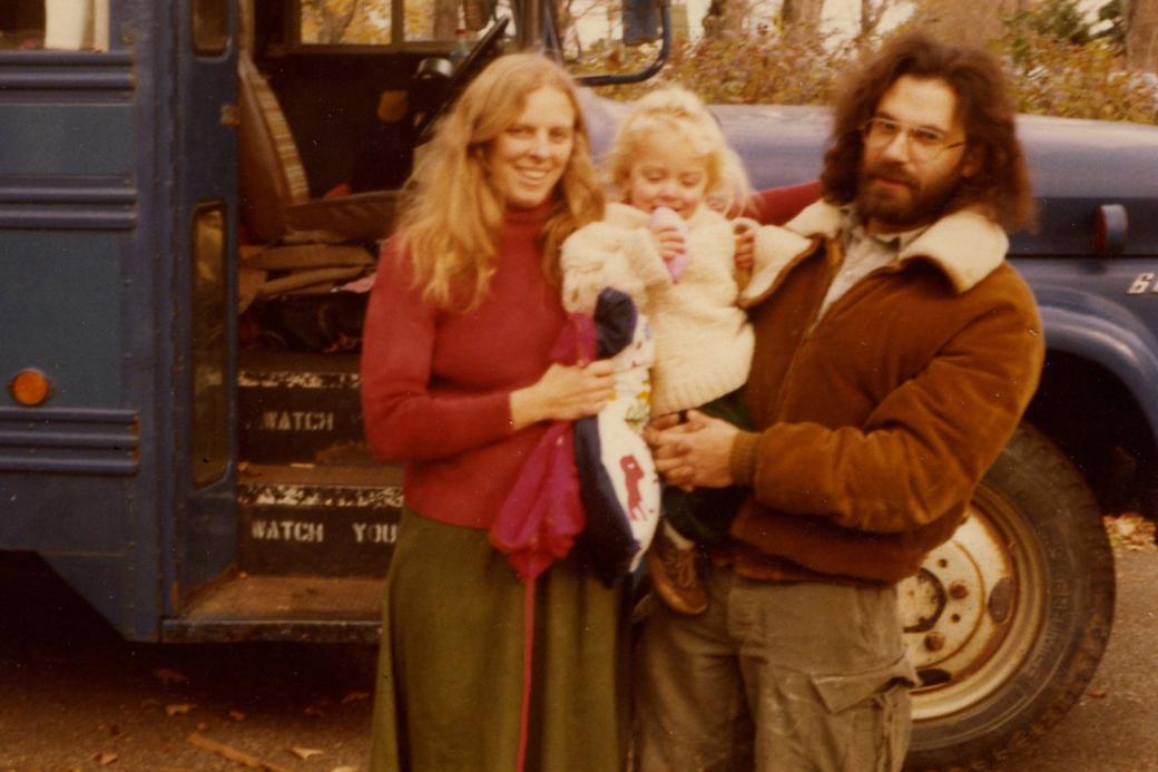 A white woman and man hold a toddler between them. The woman has blond hair and smiles at the camera, the man has long brown hair and a beard and looks unsmiling at the camera. The toddler looks downwards. In the background is a vintage blue school bus.