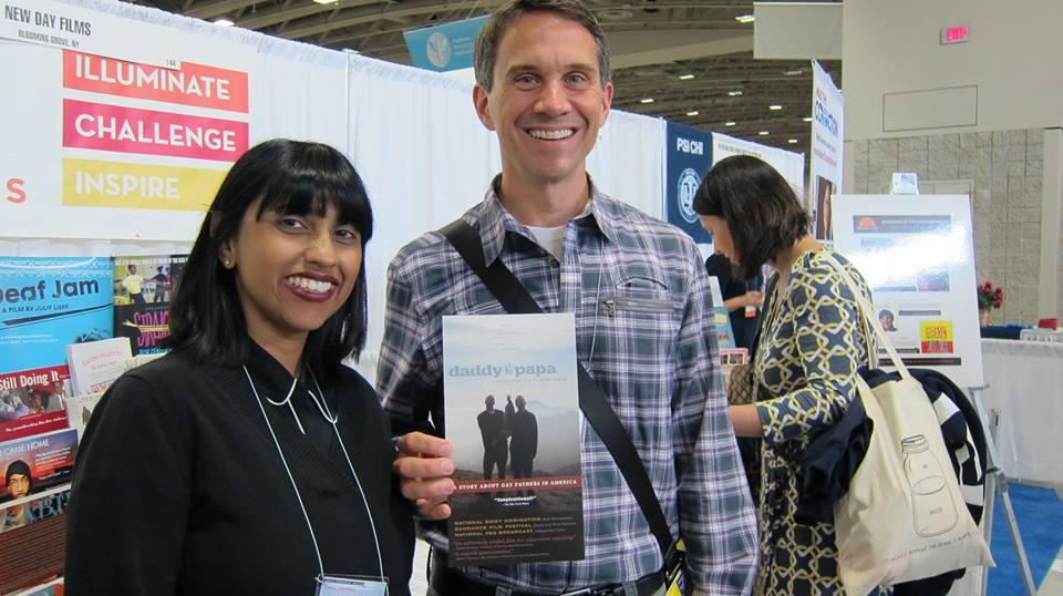 New Day filmmaker Leena Jayaswal and a man in a plaid shirt stand side by side at the New Day Films booth smiling broadly. The man holds a poster for Daddy & Papa, a film where he met his husband at a screening 12 years ago.