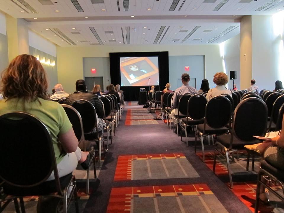 A large presentation room with attendees interspersed throughout the rows of chairs. They watch a film being screened at the front. On the screen, an image of a wooden frame with a woman’s picture in it.