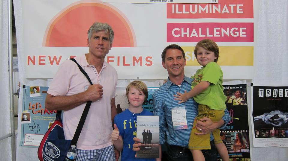 A family of two fathers and two children smile in front of the New Day Films booth. One child gives a thumbs up. One father holds up a poster for “Daddy & Papa" where he met his husband 12 years ago.