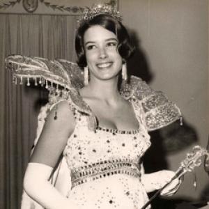 A black and white still from the New Day film By Invitation Only. A young white woman wears an elaborate, jewel encrusted dress with a large jeweled fan collar, long white evening gloves, large dangly earrings and a delicate tiara. She holds a scepter in her hands and smiles winningly as she glances off camera to the right. A curtain hangs behind her.