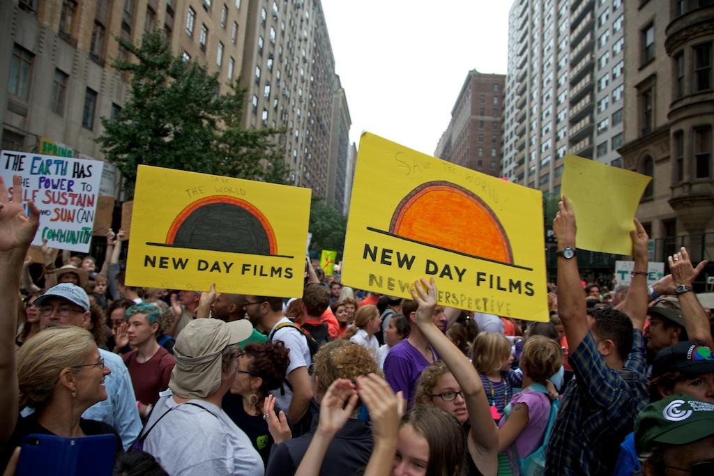 A large group of people of various races, ethnicities and genders mill around in a city street at the People’s Climate March. Several people hold posters up, three of which are yellow films with the New Day Films logo on them. Tall city buildings rise up on either side of the frame.