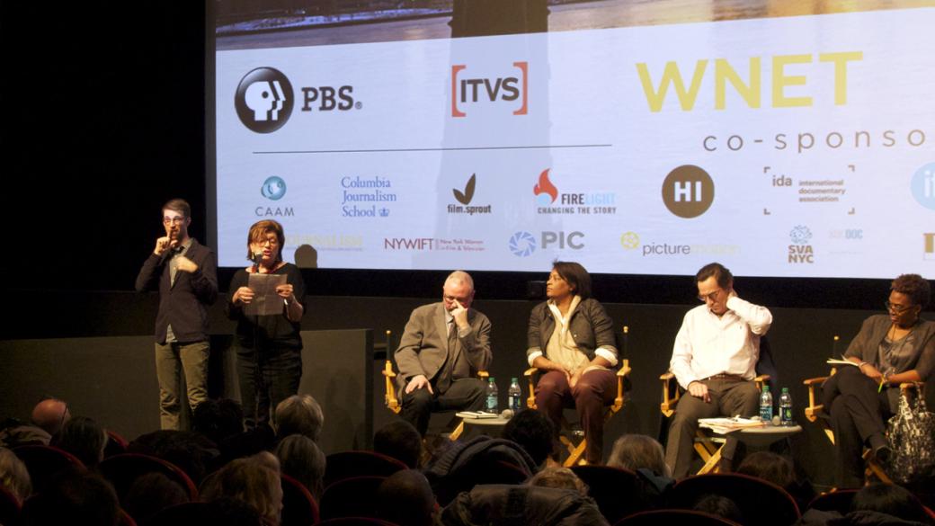 New Day filmmaker Tami Gold stands on stage in front of a screen with several sponsors’ logos on it. She is holding a microphone and reading from a paper that she is holding with her other hand. To her right is a person interpreting her words into sign language. To her left sit two of the panel members. One presses his forefinger to his temple in concentration. The other panel member looks attentively over at Tami with her hands in her lap.