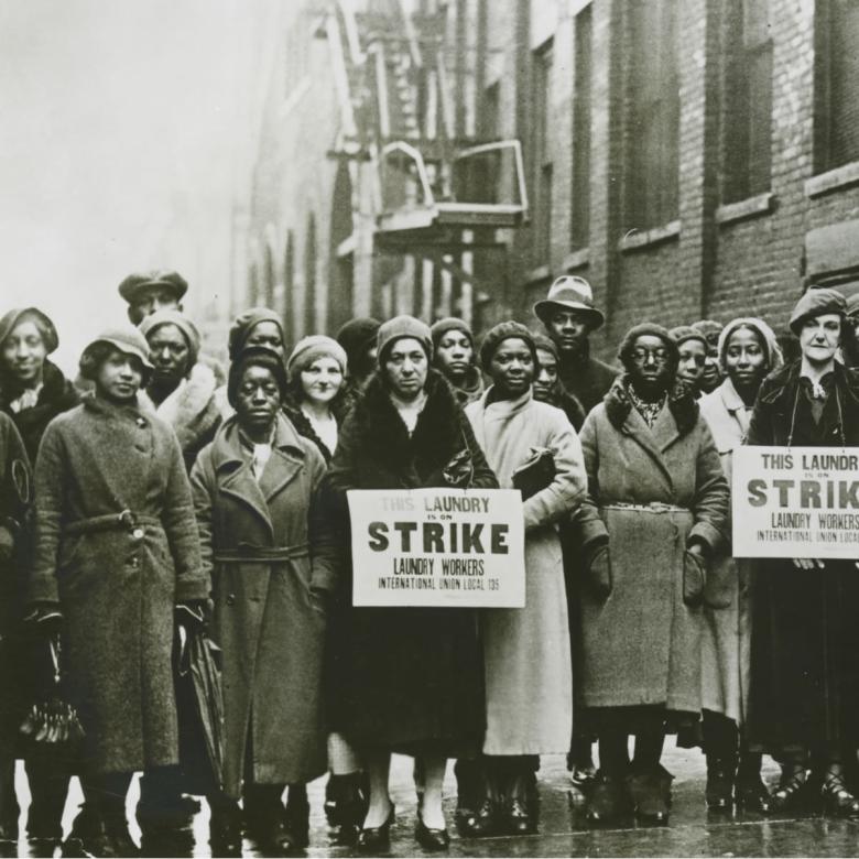 A black and white photo taken in the 1930’s of a group of striking workers who are predominantly Black women. Two women hold signs that say This Laundry is on Strike. Laundry Workers International Union Local 135.