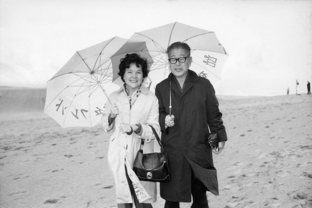 A middle-aged Japanese couple from 95 and 6 to Go stand shoulder to shoulder on the beach on a windy day. They both hold umbrellas up and smile. She wears a pale trench coat, and he waears a black trench coat.