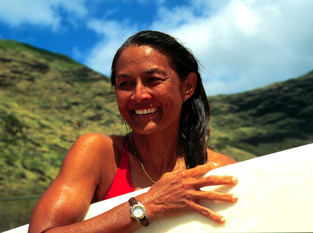 Rell Sunn, a Hawaiian woman, stands outside against a backdrop of lush green mountains and a blue sky with wispy clouds. She is wet with ocean water, her long hair pulled over to the right and draped over her shoulder. She holds a surfboard under her left arm and up against her body and smiles as she looks out to the distance.