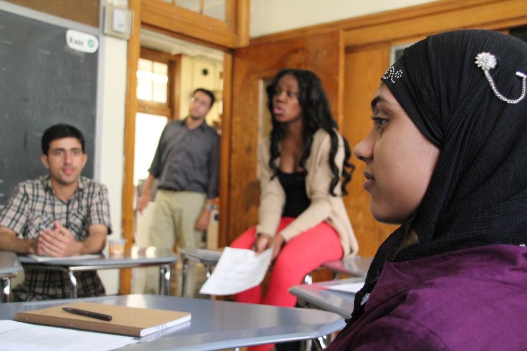 A still from the New Day film I Learn America. A profile of a teenage girl sitting in a classroom wearing a purple shirt and a black head covering. A woman in professional clothing sits on a desk to her right. She holds a piece of paper in her hand and speaks to someone out of the shot. In the background, a man wearing khakis and a button up shirt leans against the wooden door frame. All three are looking at something off camera.