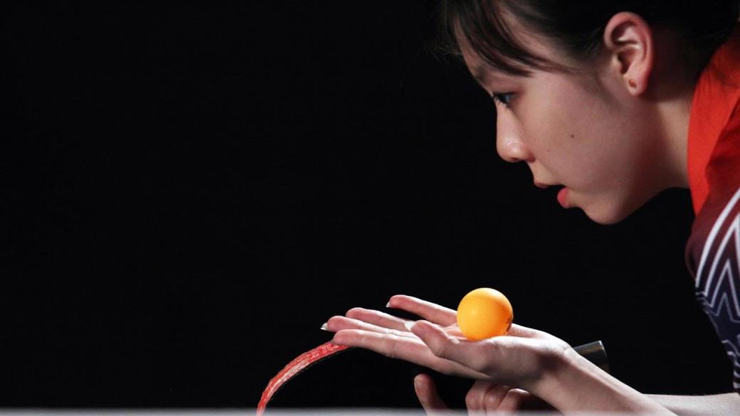 A still from the New Day film Top Spin. An orange table tennis ball rests in Teenage table tennis player Lily Zhang’s open palm. She crouches and looks in the direction of the ball, mouth slightly open in concentration. Her table tennis paddle is visible in her other hand as she prepares to serve the ball.