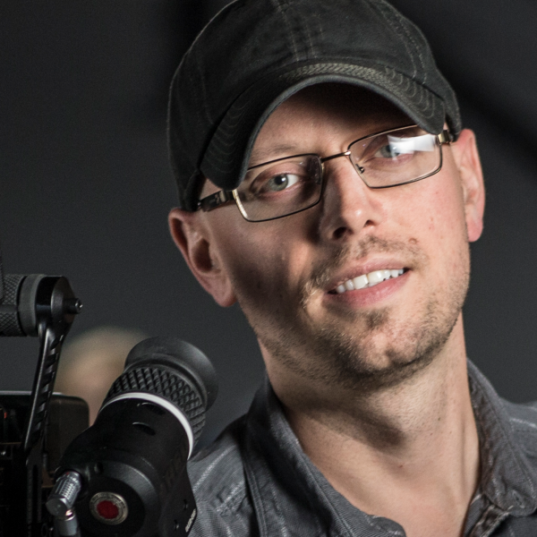 A close-up shot of Adrian's face, wearing a black baseball cap and black button-up shirt, holding a camera off to the side of the frame. 