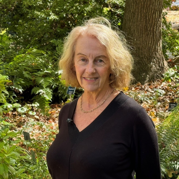 Joanne, a woman with bright blond hair and wearing a v-neck black blouse, stands in a wooded area, smiling at the camera. 