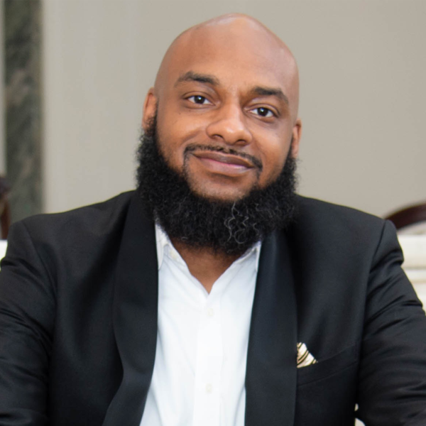 Dewey A. Ortiz, a Black man with a mid-length dark beard and slim facial hair, smiles directly at the camera. He sits in a chair, leaning forward slightly with his hands crossed in front of him. Fashionably dressed, he wears a suit jacket and a white button-up.