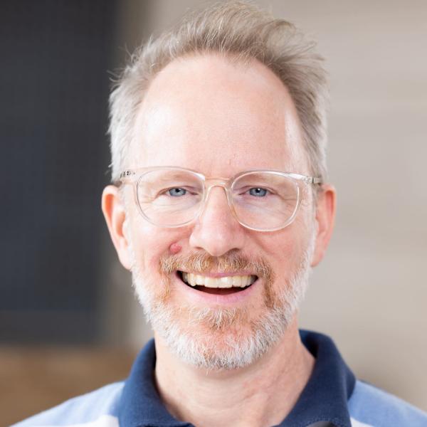 Kyle Henry is smiling, wearing glasses and a blue striped pholo shirt. 