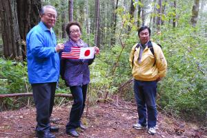 Promotional still. Three members of Fujita family display an American and Japanese flag at site of peace tree.