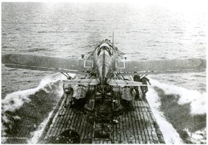 Production still. A collapsible plane sits on the deck of a submarine.