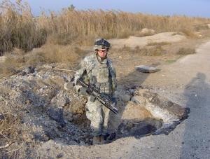 American soldier in combat gear stands in big hole created by IED explosion