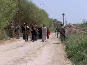 A group of Iraqi children and young men walk past armed American soldiers on a rural road