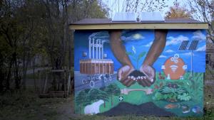 Stll from How To Power A City shows a mural on a small building. Two brown-skin hands cup a seedling over a lush food garden with solar panels on a post. Nearby, a Ford factory churns thick, gray smoke from its tall smokestacks into the jewel-toned blue sky.