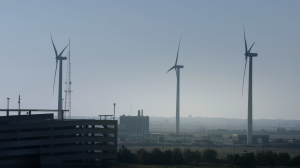 Still from How To Power A City shows three wind turbines on their very tall, slender posts towering above low buildings on a hazy day.