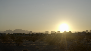 Still from How To Power A City shows the sun just rising over a city with low buildings and palm trees. It burns white-hot with a golden ring around it.