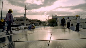 Still from How To Power A City shows workers in hard hats on a rooftop installing an array of solar panels on a cloudy day.