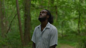 Black farmer and timber frame builder, Blain Snipstal, stands among trees on his Black Dirt Farm Collective's land