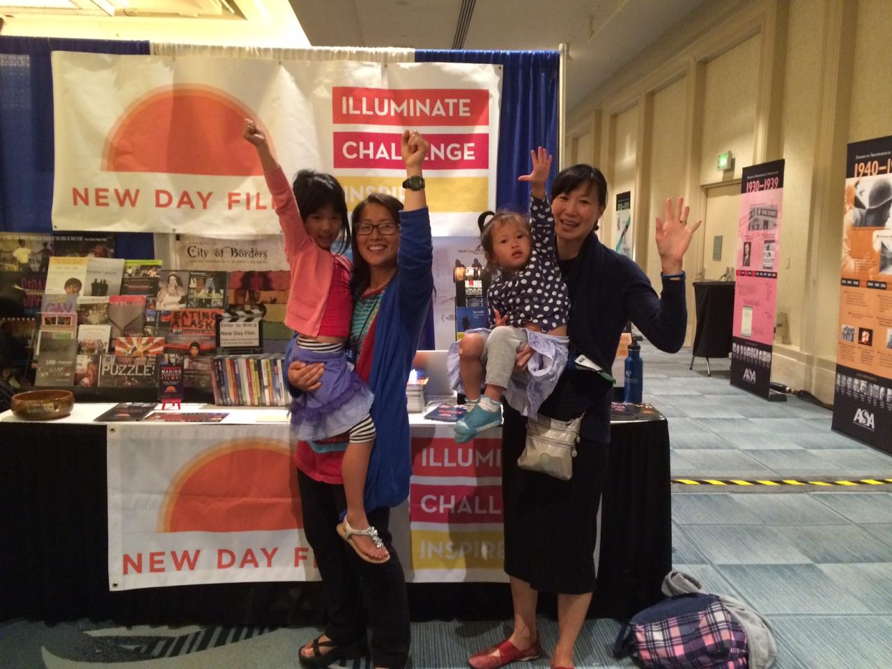 New Day Filmmakers Yun Suh and Debbie Lum each hold a young child on their hip at the New Day Films booth in the exhibition hall. All four smile and raise one fist or open hand above them in celebration.