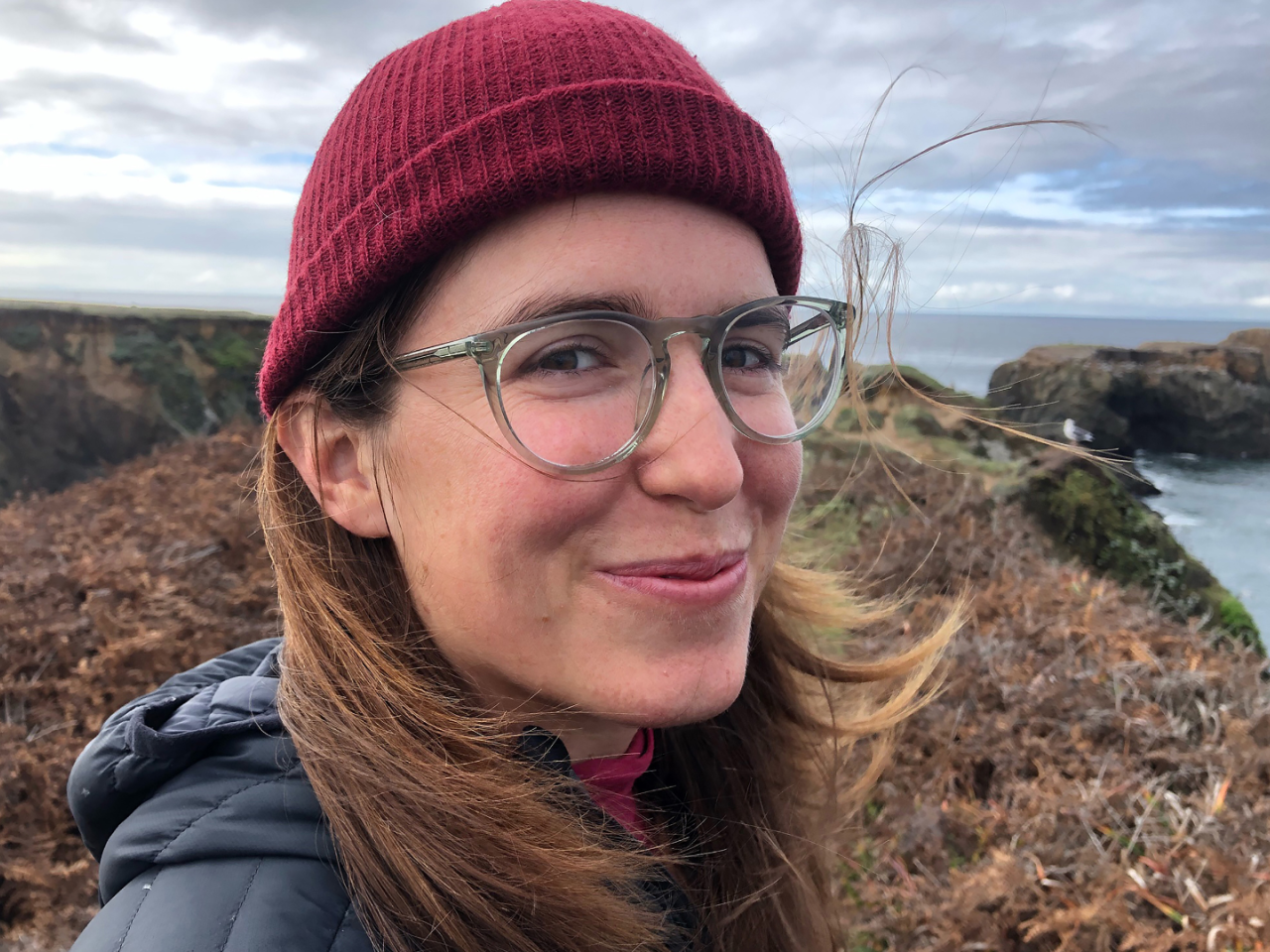 A person with long hair and glasses and wearing a red hat, looks into the camera and smiles. They are outdoors.