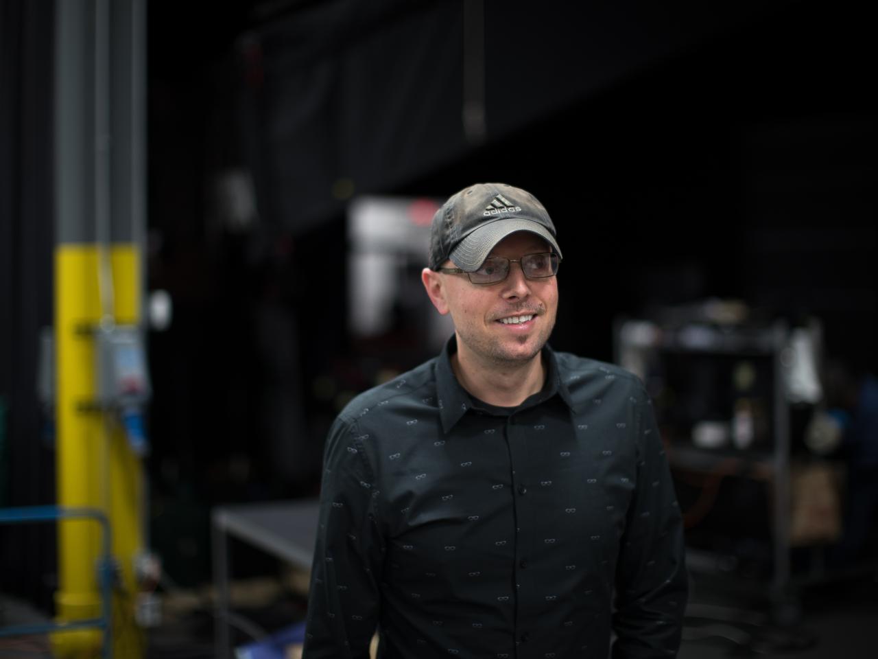 A medium shot of filmmaker Adrian Filmadria, who is wearing a black button down shirt and a baseball cap. He is looking off to the right.