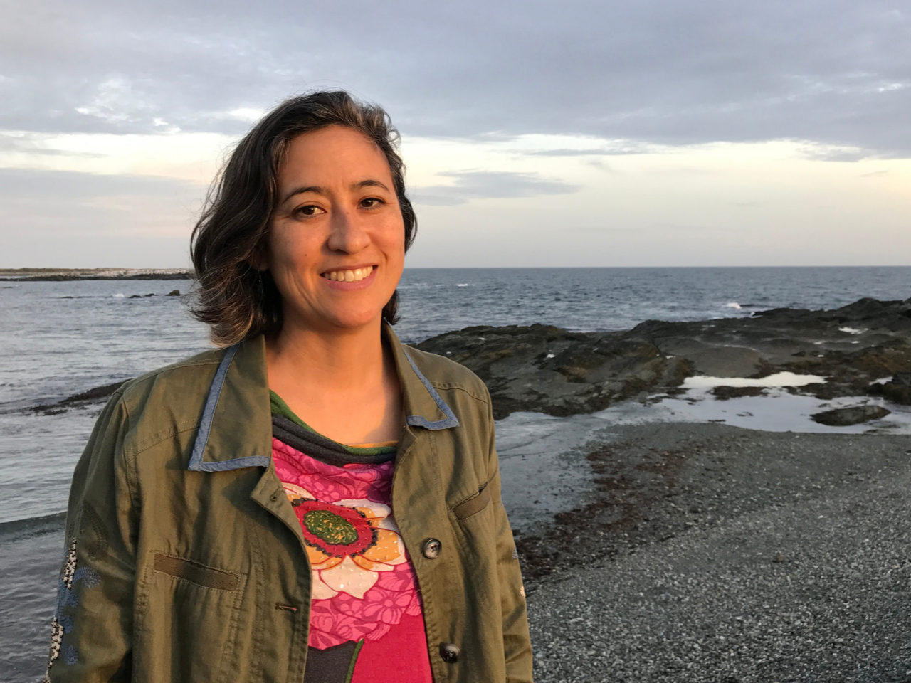 Filmmaker Julie Mallozzi is smiling on the beach with the sun on her face. She wears a dark green jacket over a colorful shirt.