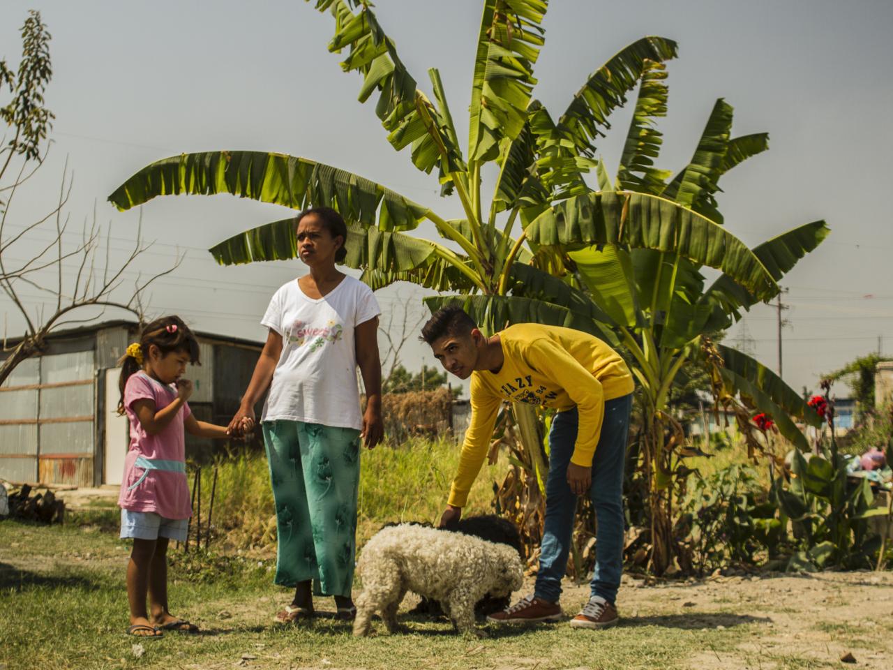 Production Still from Children of Las Brisas. Two adults and a child stand behind a tree in Venezuela. One bends to pet a small dog.