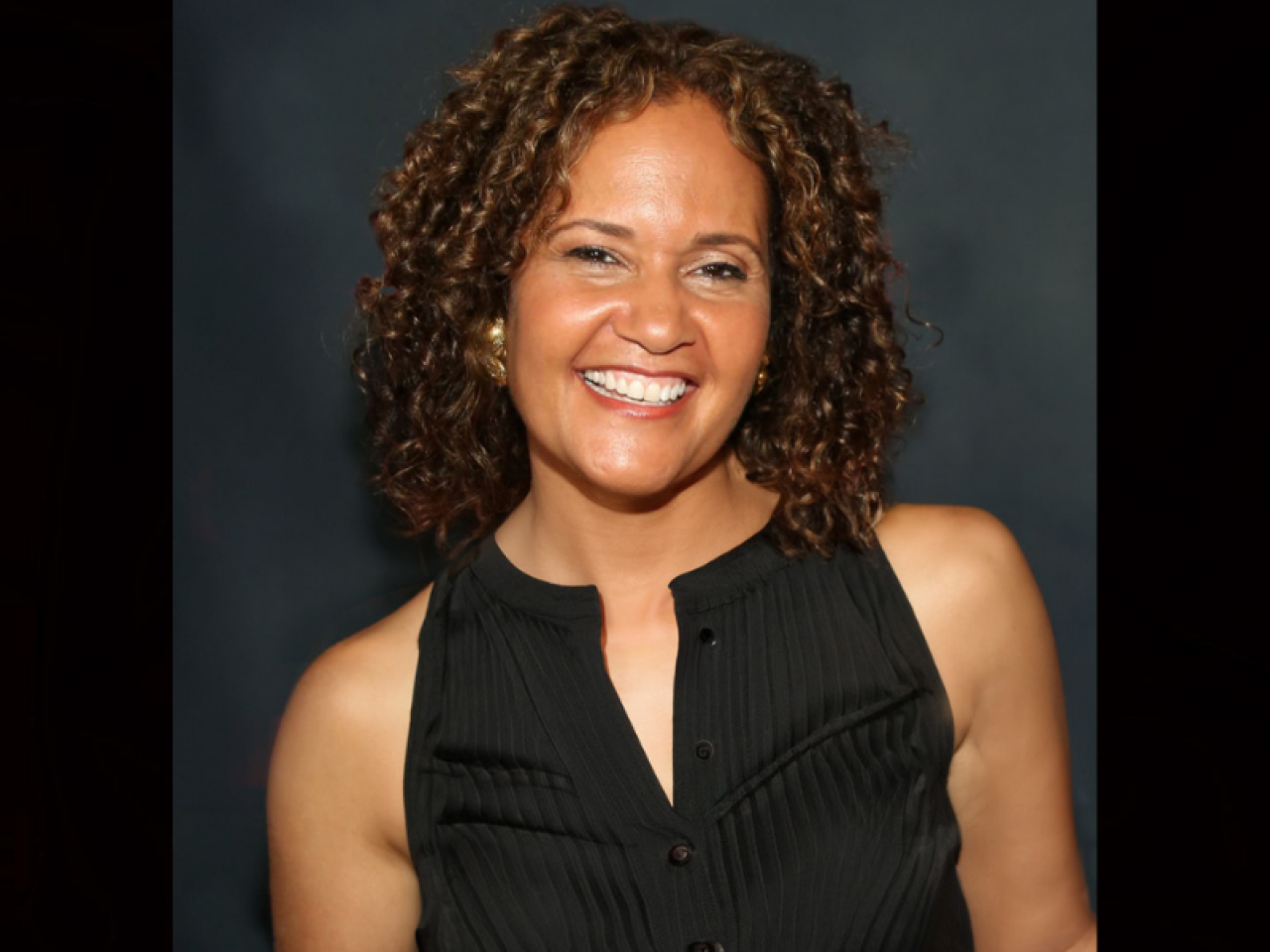 Image of a woman with dark curly hair standing in front of a navy blue background smiling broadly at the camera she wears a black tank top. 