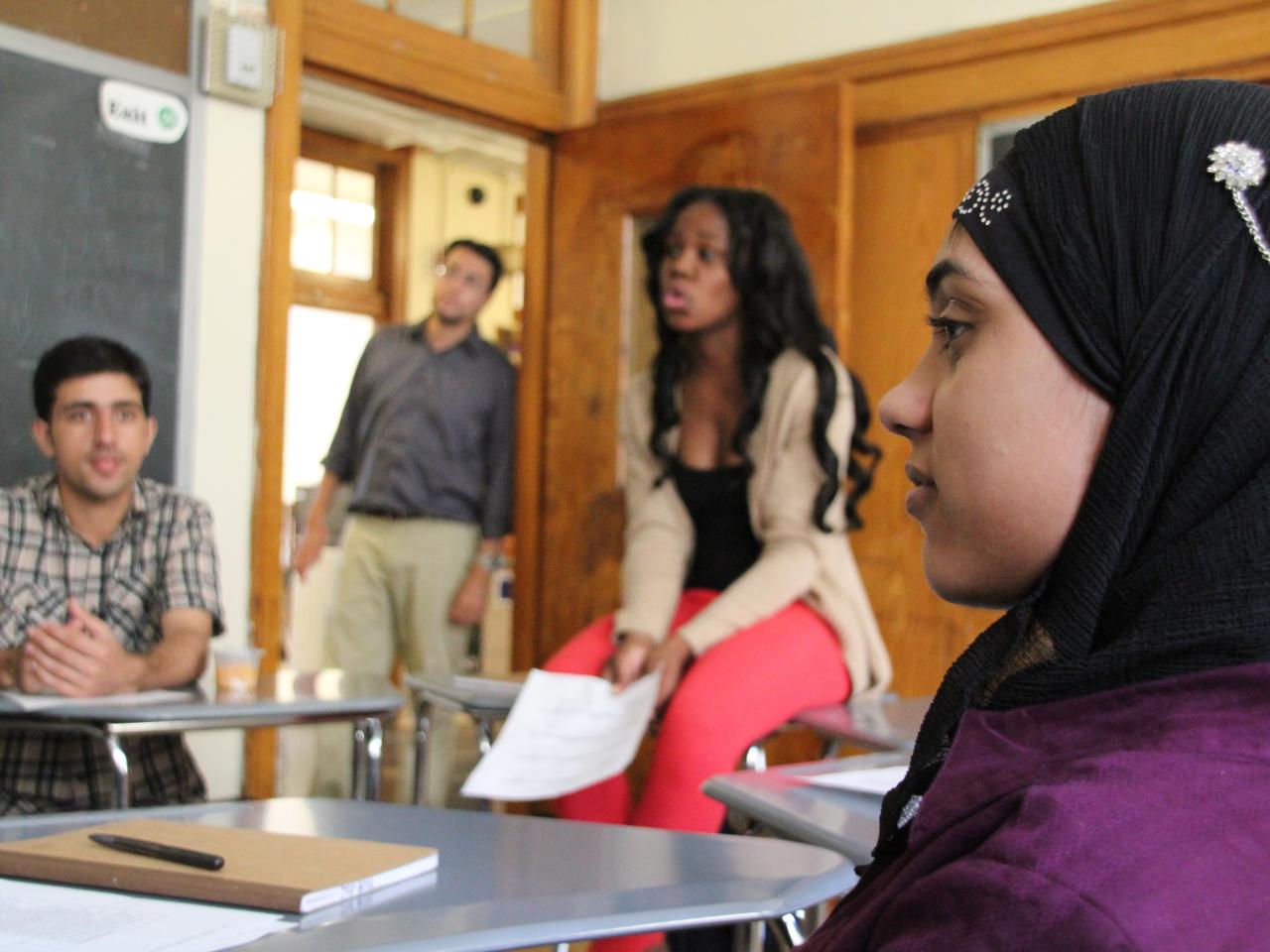 A profile of a teenage girl sitting in a classroom wearing a purple shirt and a black head covering. A woman in professional clothing sits on a desk to her right. She holds a piece of paper in her hand and speaks to someone out of the shot.