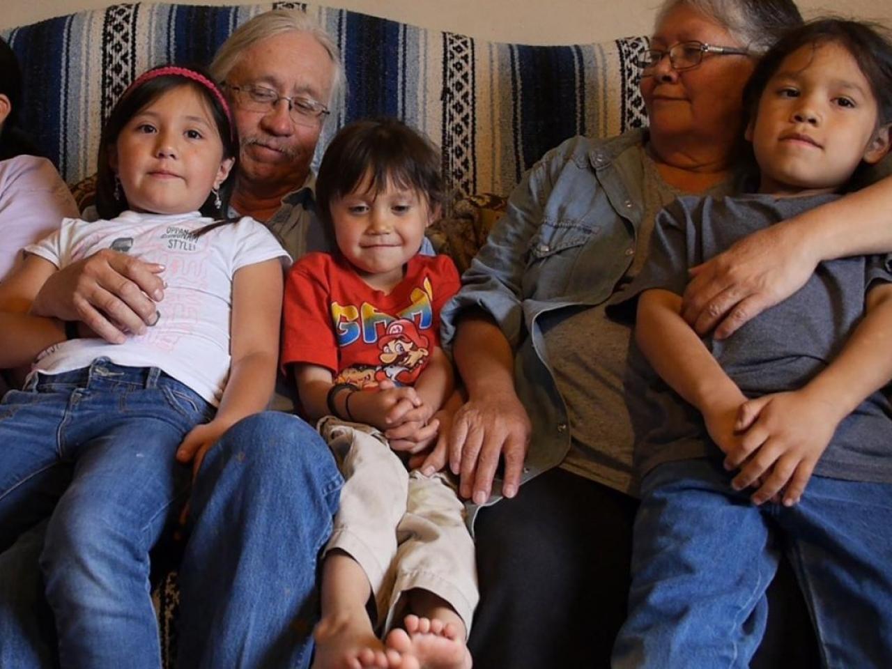 Seven members of a Blackfeet (Pikuni) family, the Mombergs, sit together on a sofa ranging from elders to pre-school age. The youngest three children sit on their grandparents’ laps and look ahead or down while the two older youth smile at something off in the distance.