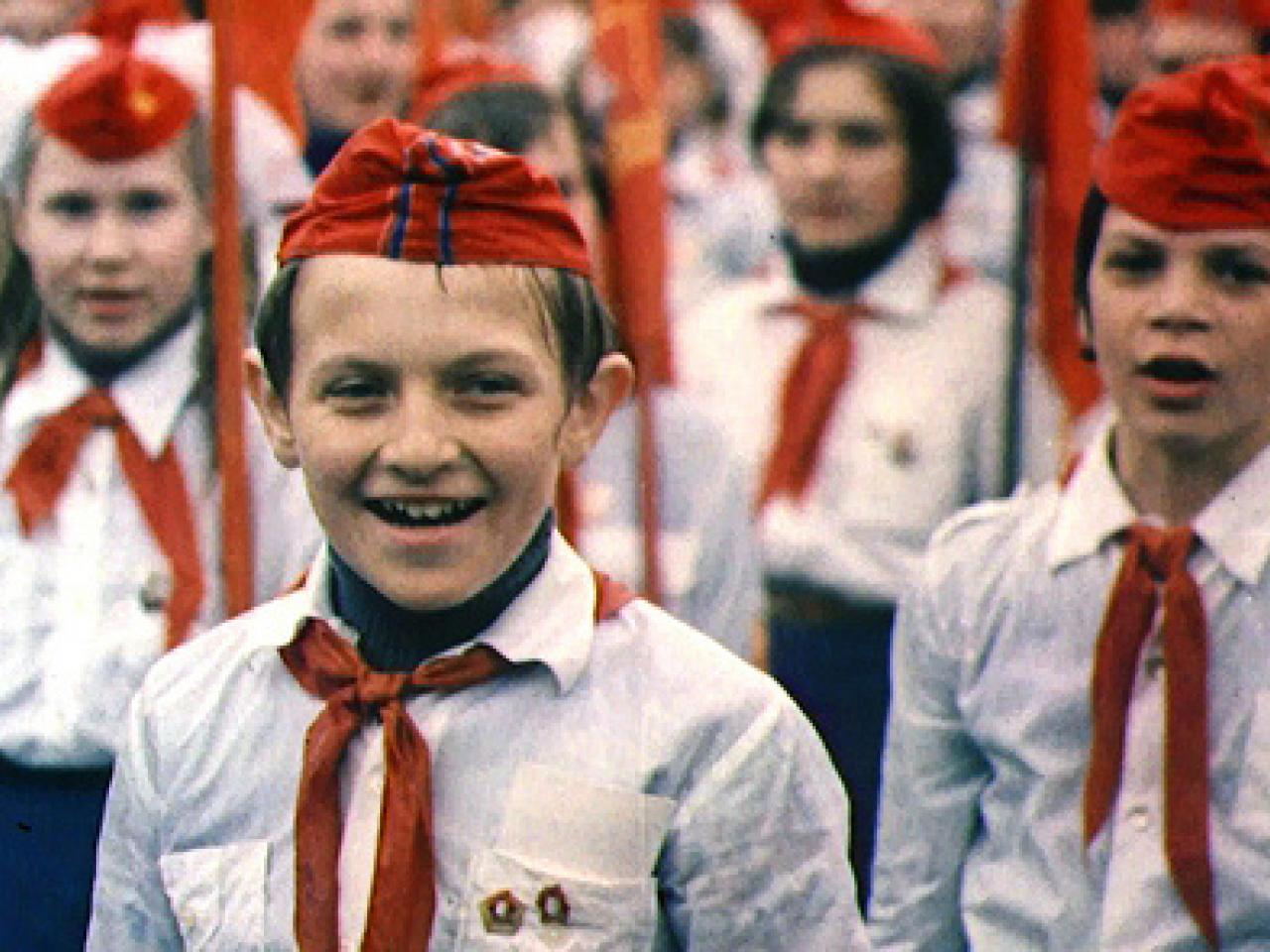 Russian Children stand on guard wearing military outfits which include red hats and red scarves, white shirts with pins on their pockets.